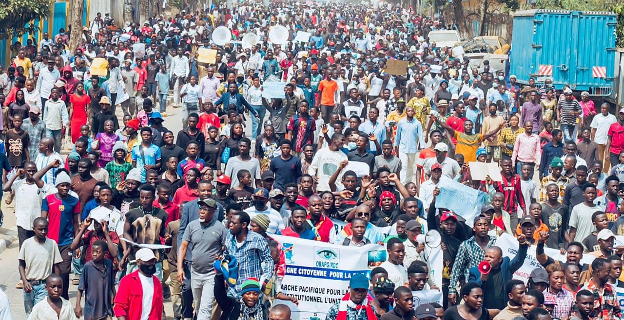 A dense crowd of demonstrators marches down an avenue behind a civil society banner